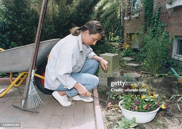 Eine junge Frau jätet im Garten Unkraut mit einer Kombihacke. Neben ihr steht eine Schubkarre, an die eine Harke und ein Rechen angelehnt sind....