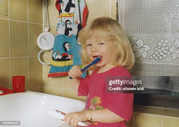Mit Zahnbürste und Zahncreme ausgestattet putzt sich ein kleines Mädchen im Badezimmer ihre Zähne. Auf dem Waschbeckenrand steht ein Zahnputzbecher...
