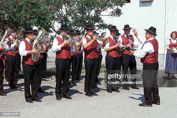 Musiker mit Trachtenweste und Hut blasen auf ihren Blechblasinstrumenten. Vor den Bläsern der Dirigent. Blasmusik; Kapelle; musizieren; Instrument;...