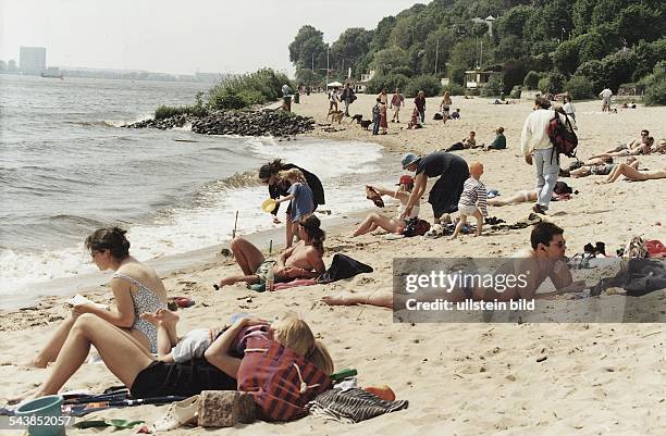 Menschen in der Sonne am Strand bei Övelgönne an der Elbe. Die Strandgäste lesen, liegen, stehen, gehen, ziehen sich an oder aus oder um, tragen...