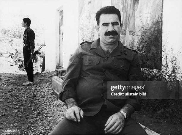 Der Vorsitzende der PKK Abdullah Öcalan sitzt vor einem Gebäude. Im Hintergrund steht ein Mann mit einem Gewehr. Aufgenommen September 1991.