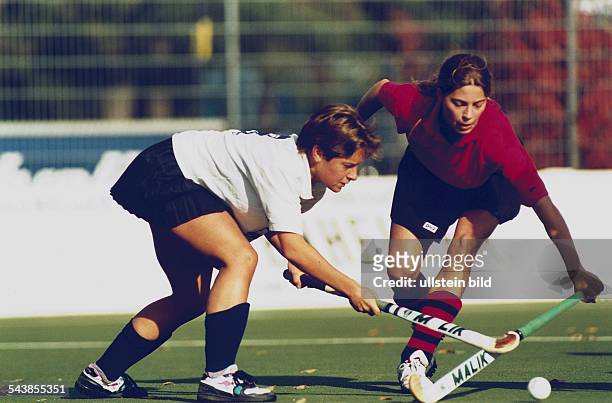 Feldhockey: Spielerinnen zweier Mannschaften kreuzen auf dem Spielfeld ihre Schläger, während der Ball auf dem Rasen auf sie zurollt. Aufgenommen um...