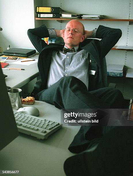 Ein Mann hat an seinem Computerarbeitsplatz die Füße auf den Schreibtisch gelegt und schläft. Mit im Nacken verschränkten Armen hält er seinen Kopf....