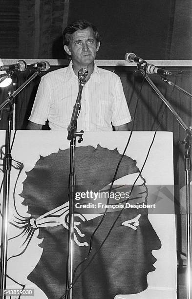 Union of the Corsican People leader Edmond Simeoni stands at a podium which displays the Corsican flag during a press conference.