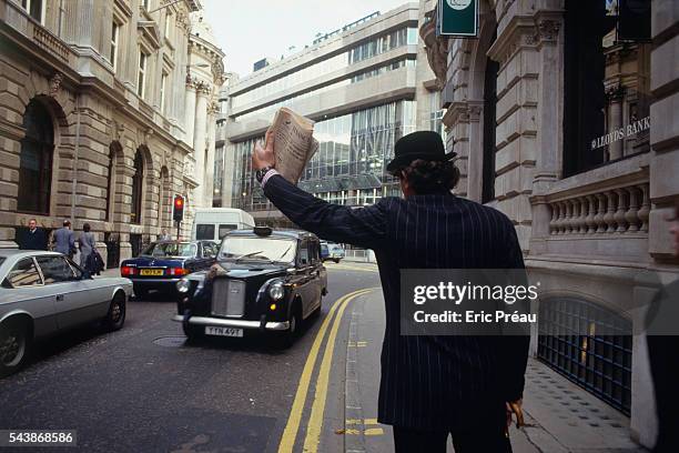 An English businessman in a three-piece suit and bowler hat, or derby, holds an umbrella and waves the Financial Times to hail a cab in front of the...