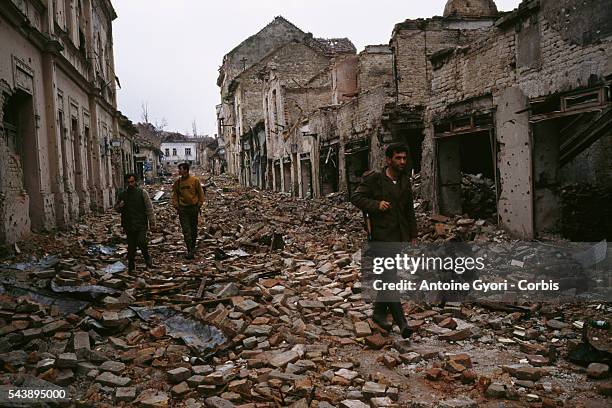 Yugoslavian soldiers and Serb paramilitaries, including Zeljko "Arkan" Raznatovic, walk past bombed buildings riddled with bullet holes and streets...