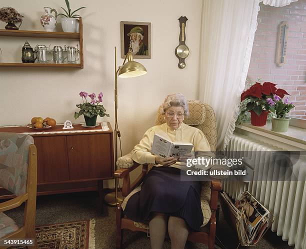 Wohnen im Alter - eine aeltere Frau sitzt in ihrem Wohnzimmer im Sessel und liest ein Buch