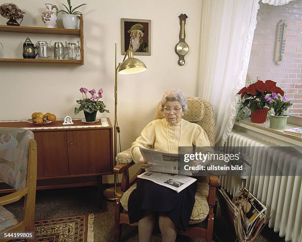 Wohnen im Alter - eine aeltere Frau sitzt in ihrem Wohnzimmer im Sessel und liest eine Zeitung