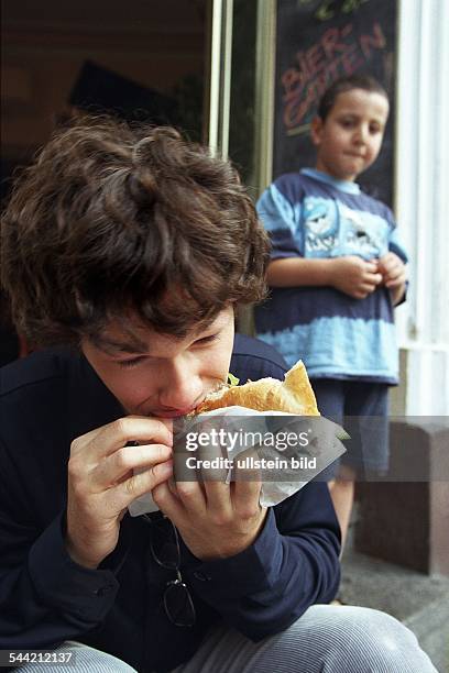 Deutschland: Fastfood, hier: Doener, Student isst Doener auf der Strasse und ein kleiner Junge schaut zu.