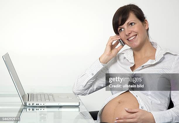 Schwangerschaft, schwangere junge Frau am Schreibtisch telefoniert mit dem Handy, auf dem Schreibtisch ein Laptop