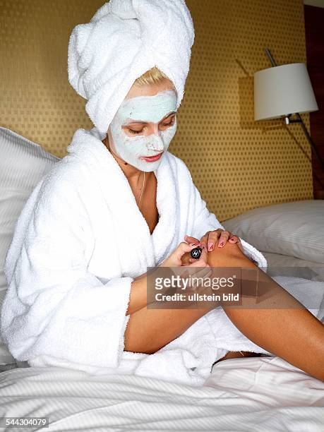 Kosmetik, Wellness, Wellnesswochenende, Wellnesshotel, Frau im Bademantel auf dem Bett mit Hantuchturban, Gesichtsmaske lackiert sich die Fingernaegel