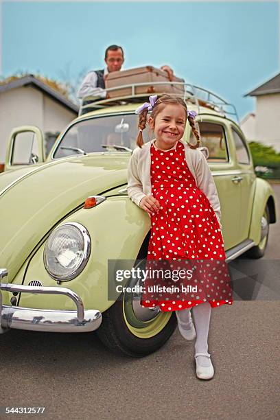 Familienfoto im Stil der 50er-Jahre. Der Volkswagen Baujahr 1955 steht im Mittelpunkt. Szene: Blondes Mädchen mit Zöpfen posiert vor dem Auto,...