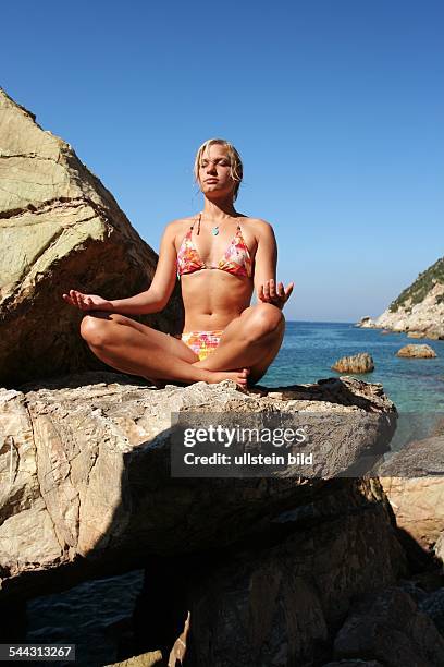 Italien, Urlaub auf Elba. Junge Frau im Bikini entspannt sich bei einer Yogauebung auf einem Felsen am Meer.