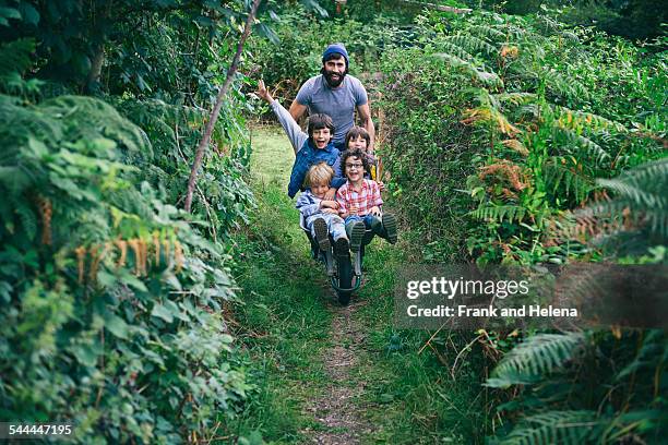 mid adult man pushing wheelbarrow full of boys in garden - summer press day ストックフォトと画像