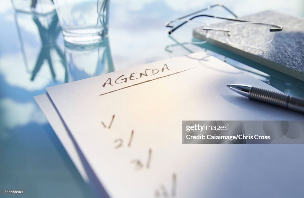 Close up of desk with,glass,glasses and note saying agenda,pen