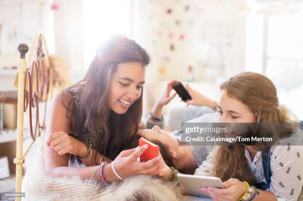 Three teenage girls using electronic devices while lying on bed