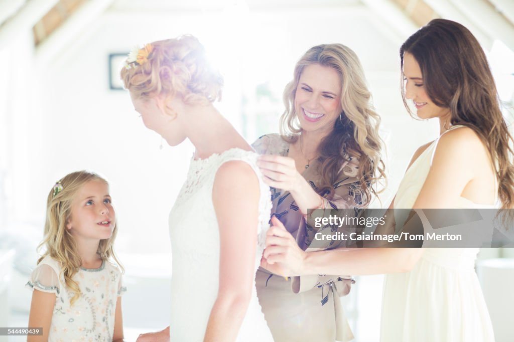 Matron of honor and bridesmaid helping bride with dressing in domestic room