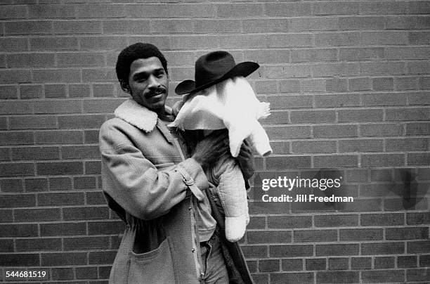 Man holding a baby, whose head is covered with a blanket and a cowboy hat, USA, circa 1975.