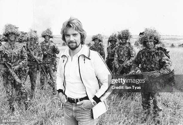 Presenter Noel Edmonds with a group of soldiers in camouflage, whilst filming a television show on Salisbury Plains, England, 1976.