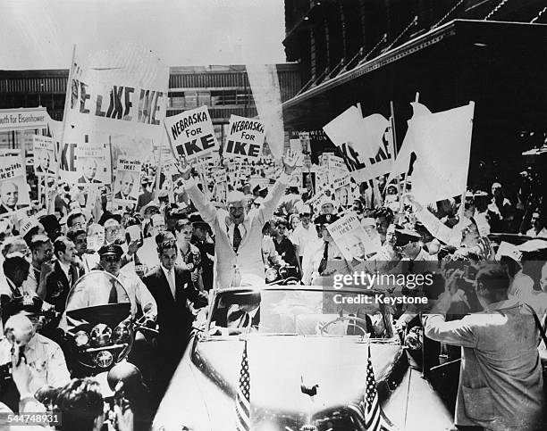 General Dwight Eisenhower raising his arms and smiling in the back of his car, as he drives though the crowds of supporters holding 'We Like Ike'...