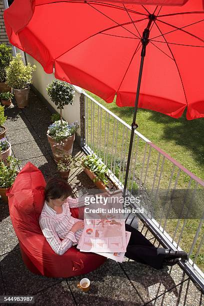 Junge Frau liest auf einem Sitzsack sitzend Zeitung auf dem Balkon, LåNEBURG, NIEDERSACHSEN, DEUTSCHLAND, . |Young woman reading newspaper on her...