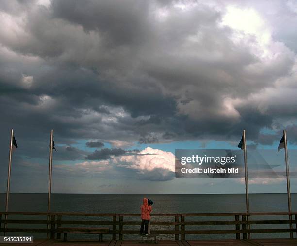 Deutschland, Mecklenburg-Vorpommern, Insel Rügen, Junge schaut vor einer Wokenlandschaft von der Seebrücke in Binz durch ein Fernrohr auf das Meer