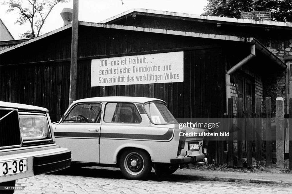 GDR, Brandenburg, Waltersdorf - Car (Trabant) in front of the sign "DDR - das ist Freitheit, sozialistische Demokratie und Souveraenitaet des werktaegigen Volkes", in the foreground the partial viewof a volvo - 1985