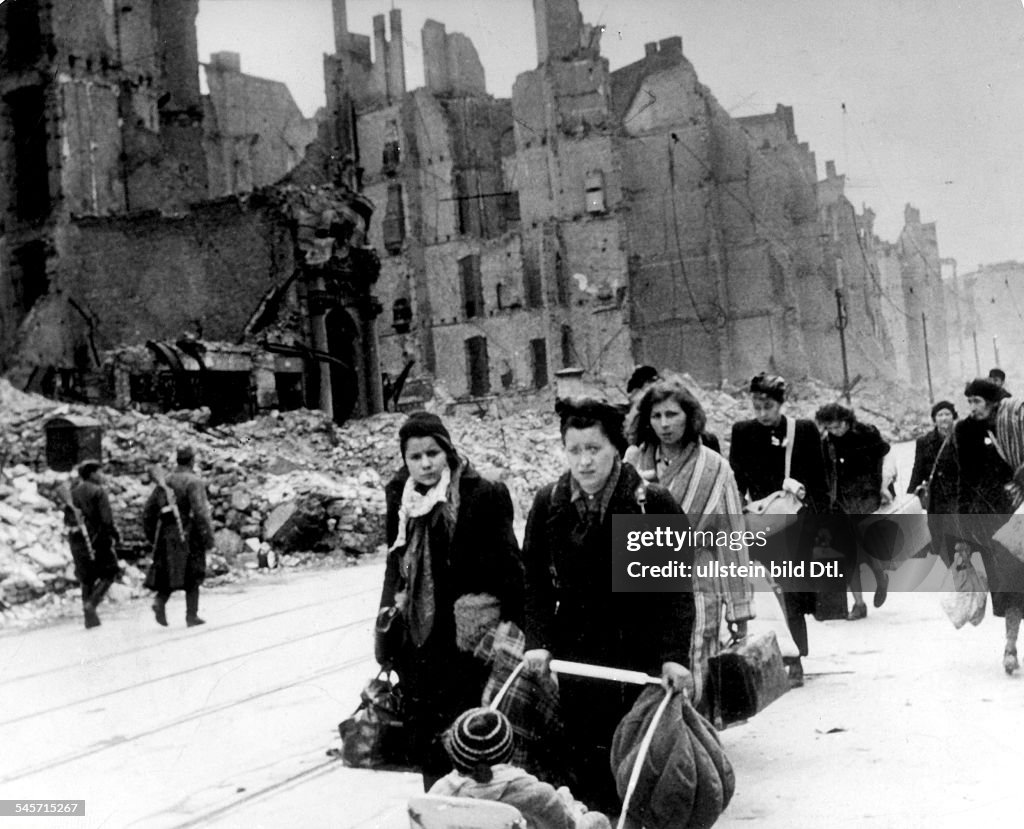 Stream of refugees and people who have been bombed out of their homes moving through destroyed streets - 1945after end of war; on the left two soviet soldiers patrolling)