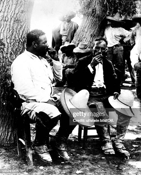 Mexican revolutionary leader Pancho Villa sits against a tree, smoking with Mexican General Porfirio Ornelas Galindo, their straw hats over their...