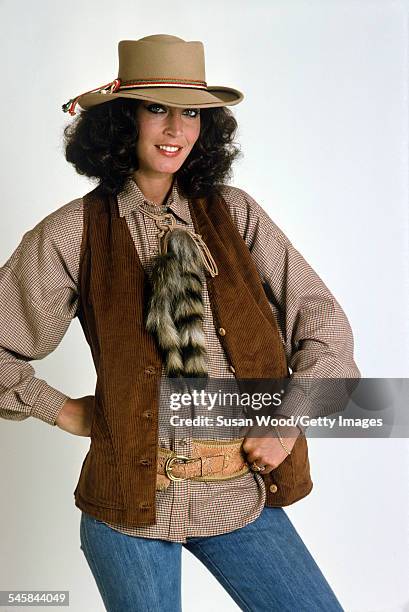 Portrait of an unidentified model, dressed in a brown plaid shirt, a striped animal tail necktie, a brown suede vest, and a creme hat, as she poses...