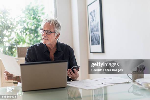 usa, new jersey, senior man sitting in home office and using laptop - business people and paper imagens e fotografias de stock