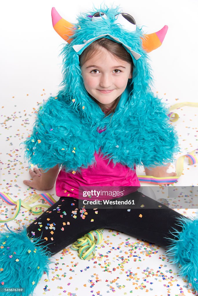 Smiling girl masquerade as a monster sitting on white ground covered with confetti