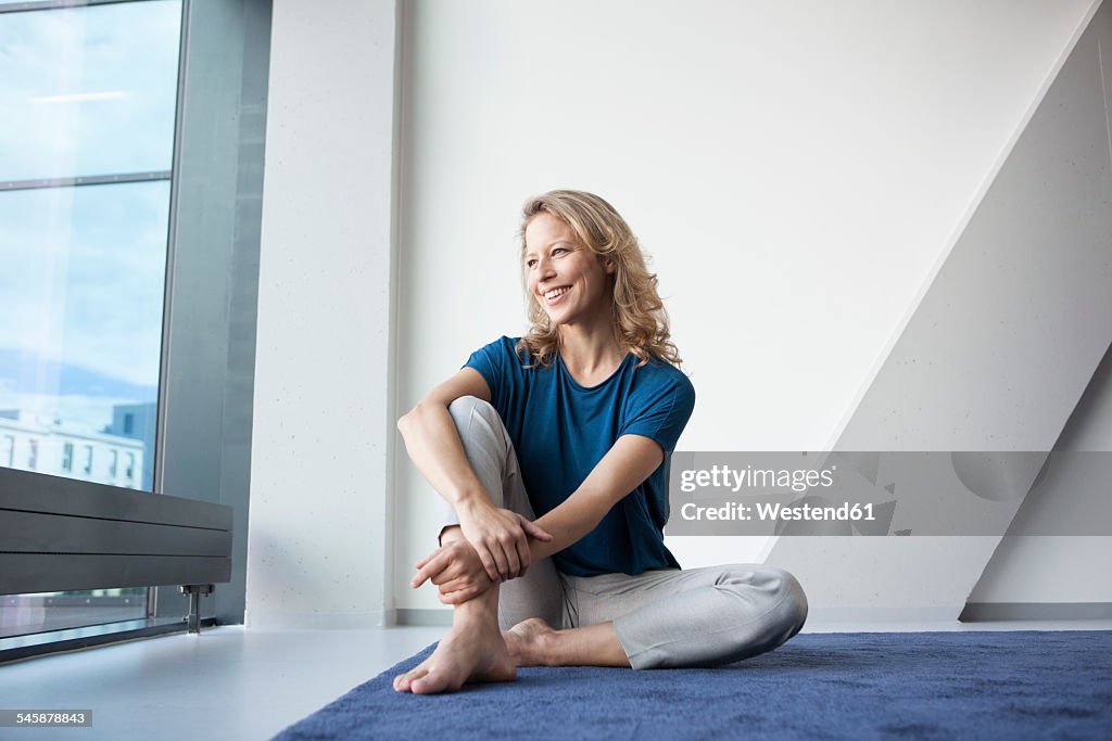 Portrait of happy mature woman sitting on carpet at home looking through window