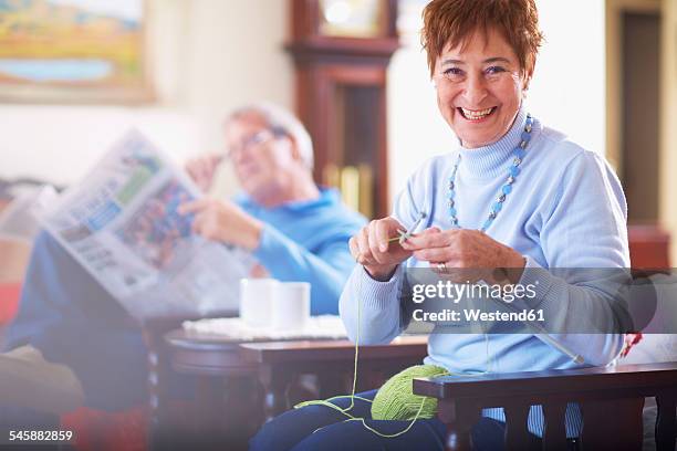 senior woman knitting with husband in background reading newspaper - stricknadel stock-fotos und bilder