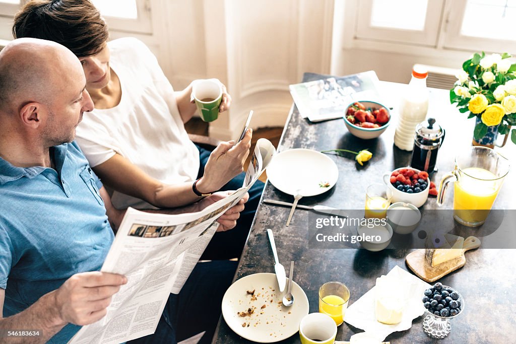 Französisches Paar mit Handy und Zeitung am Frühstückstisch