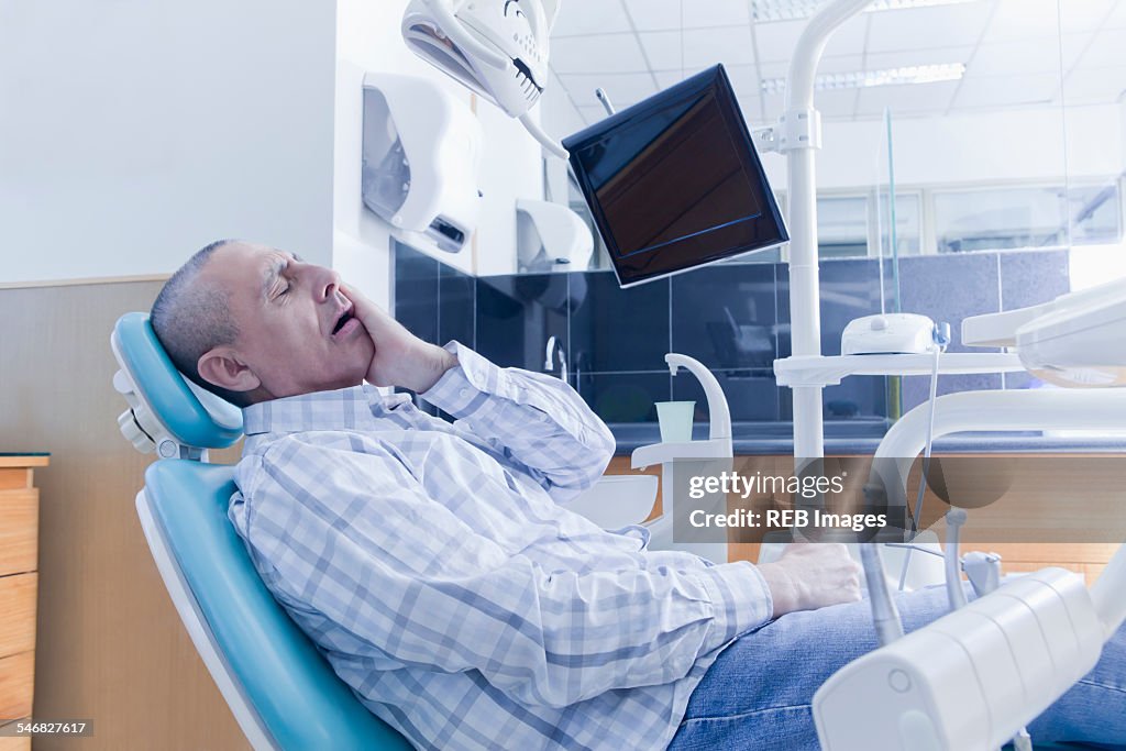 Older Hispanic man holding cheek in chair at dentist