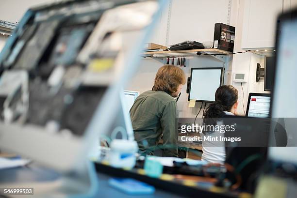 Computer Repair Shop. Two people seated using screens, and computer monitors in various stages of repair. 