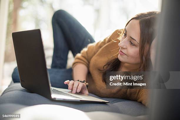 woman lying on a sofa looking at her laptop, smiling. - zurücklehnen stock-fotos und bilder