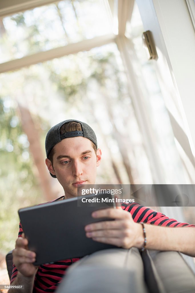 Man wearing a baseball cap backwards, sitting on a sofa, looking at a digital tablet.