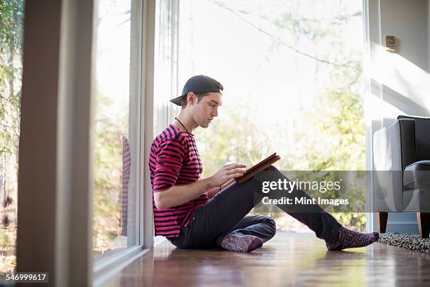 man wearing a baseball cap backwards, sitting on the floor in a living room, looking at a digital tablet. - back to front stock pictures, royalty-free photos & images