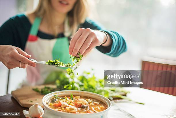 a woman wearing an apron, sitting at a table, sprinkling herbs into a bowl of vegetable stew. - vegetable soup stockfoto's en -beelden