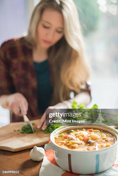 a woman sitting at a table, chopping herbs, a bowl of vegetable stew in the foreground. - chopped herbs stock pictures, royalty-free photos & images