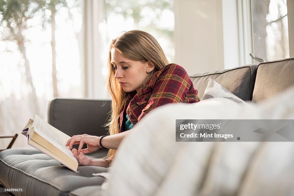 Young woman lying on a sofa, reading a book.