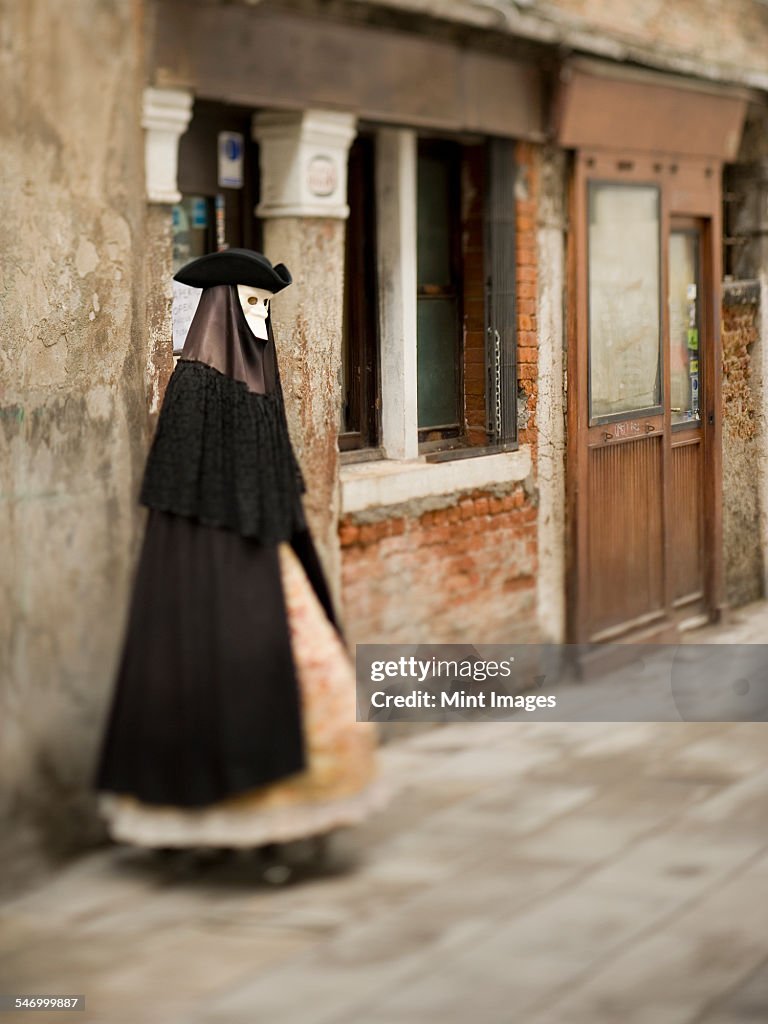 A person in carnival costume, a woman in a black cloak and dress with petticoats, wearing a white face mask and tricorn hat. 