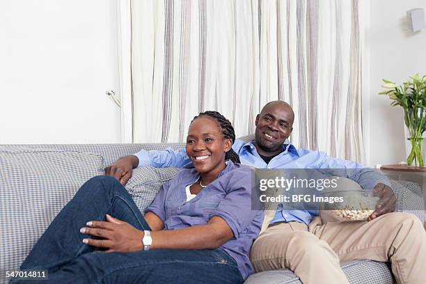 african husband and wife reclining on sofa watching television and eating popcorn , cape town, south africa - south africa women fotografías e imágenes de stock