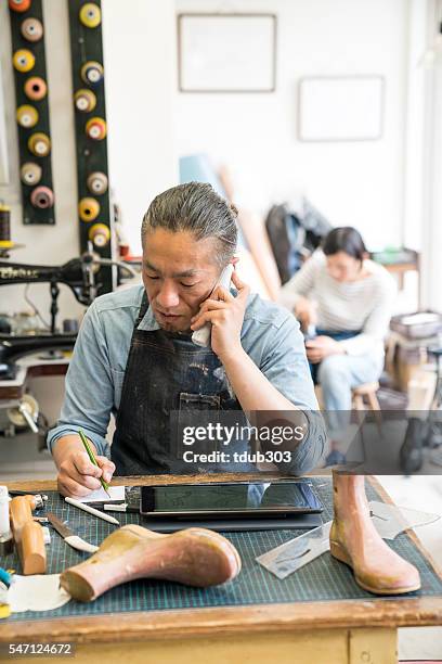 cobbler talking on a smartphone in his workshop - cobbler stock pictures, royalty-free photos & images