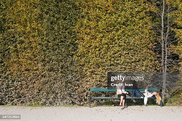 two young woman sit on a bench in a leisure time in front of green and yellow leaf wall - regents park stock pictures, royalty-free photos & images