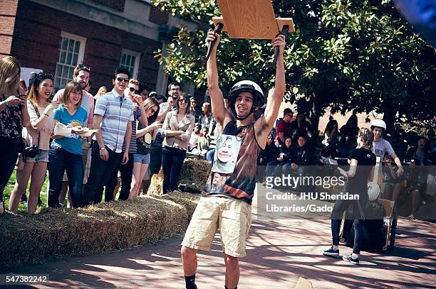 Crowd watches from the sidelines as a male student wearing tank top and helmet smiles with his arms in the air, wielding a wooden contraption,...