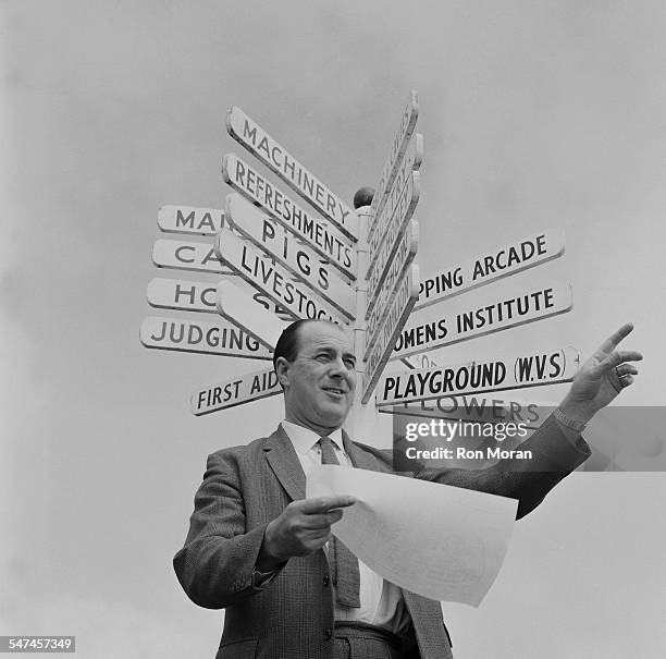 Secretary of the Royal Cornwall Show, Albert Riddle, stands beneath a sign showing events taking place at the agricultural show, circa June 1967.