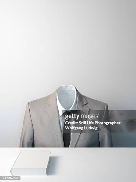 a headless body in a suit with white shirt and black tie sits in front of a white book on a plain white desk - headless stockfoto's en -beelden